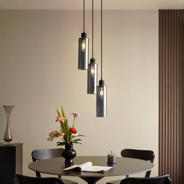 Moderne 3-flammige Pendelleuchte mit zylindrischen Rauchglasschirmen über einem dunklen runden Esstisch mit Stühlen und Blumenstrauß.