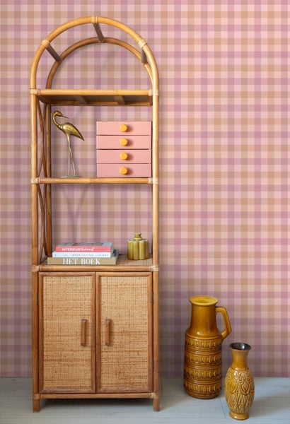 Karierte Tapete in Rosa und Beige hinter einem Standregal aus Rattan mit Dekorationsartikeln.