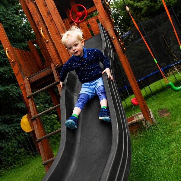 Kind rutscht auf einer dunklen Kunststoffrutsche an einem Spielturm aus Holz im Garten.