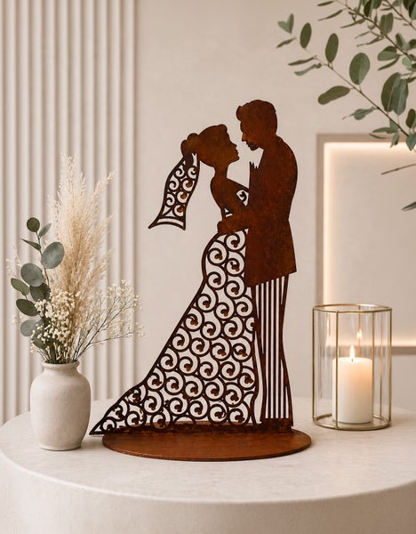 Dekorative Szene mit einer Metallfigur eines Hochzeitspaares mit filigranen Mustern auf einem Tisch, neben einer Vase mit Trockenblumen und einer Kerze.