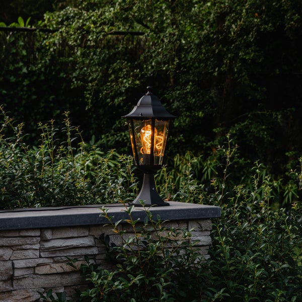 Klassische schwarze Außenlaterne mit warmem Filament-Leuchtmittel auf einer Steinmauer umgeben von grünen Pflanzen im Garten.