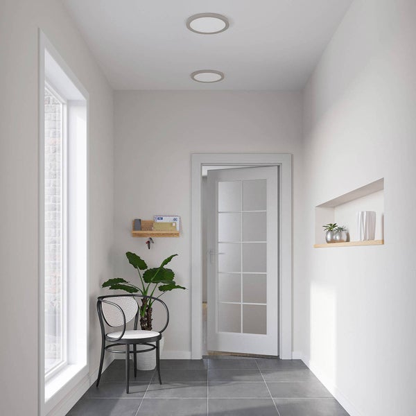 Heller Flur mit weißer Glastür, grauen Bodenfliesen, zwei runden Deckenleuchten, einem Stuhl und einer Zimmerpflanze.