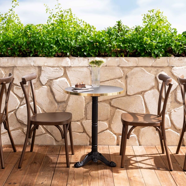 Helle Außenszene mit rundem Bistrotisch mit schwarzem Standfuß, zwei hölzernen Stühlen, einer Holzterrasse und einer Steinmauer mit grüner Hecke.