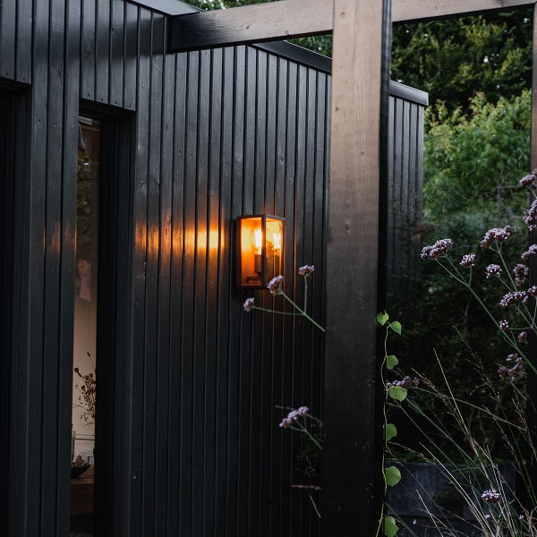 Moderne rechteckige Außenwandleuchte mit warmem Licht an einer dunklen vertikalen Holzfassade.