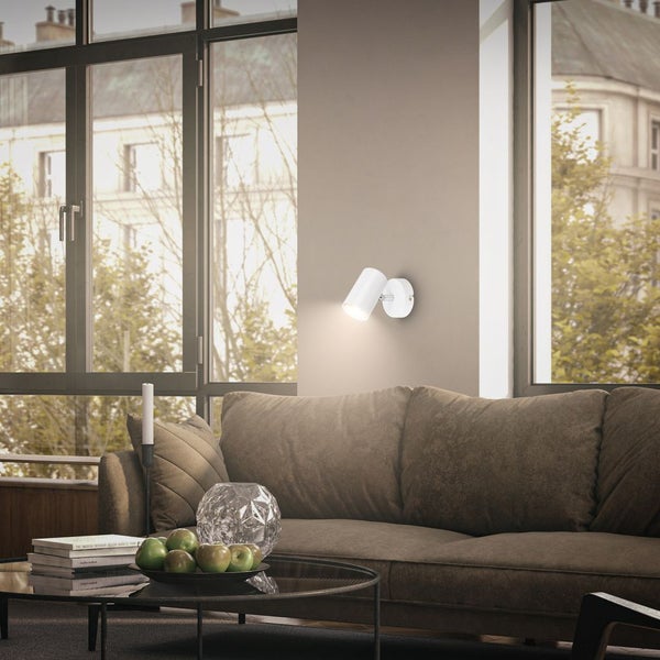 Moderner weißer Wandstrahler über einem braunen Sofa in einem hellen Wohnzimmer mit großen Fensterfronten.