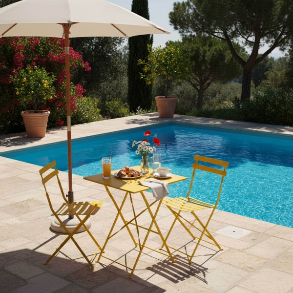 Mediterrane Terrasse am Pool mit gelber Bistrogarnitur aus Klapptisch und Stühlen unter einem großen Sonnenschirm, umgeben von einem grünen Garten.