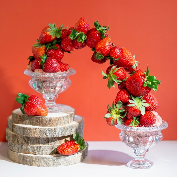 Erdbeeren-Arrangement als Bogen zwischen zwei Glasschalen auf einem Stapel aus Holzscheiben.