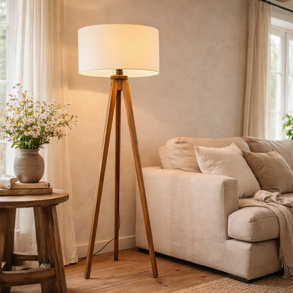 Dreibeinige Stehlampe aus Holz mit hellem Stoffschirm in einem hellen Wohnzimmer neben einem Sofa und einem Beistelltisch.