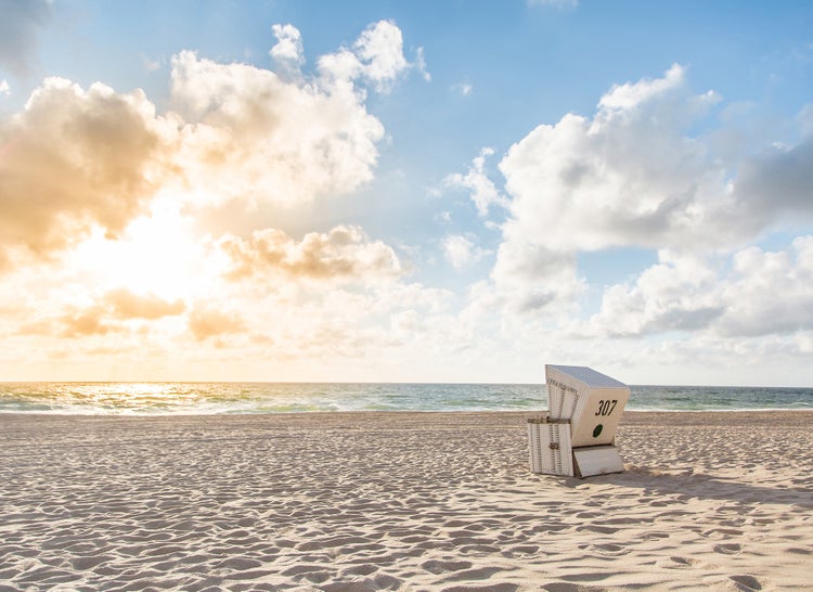 Weißer Strandkorb aus Geflecht auf einem weiten Sandstrand mit Blick auf das Meer unter einem bewölkten Himmel bei Sonnenuntergang.
