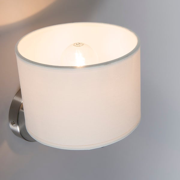 Wandleuchte mit weißem zylindrischem Stoffschirm und Metallsockel inklusive leuchtendem Leuchtmittel.