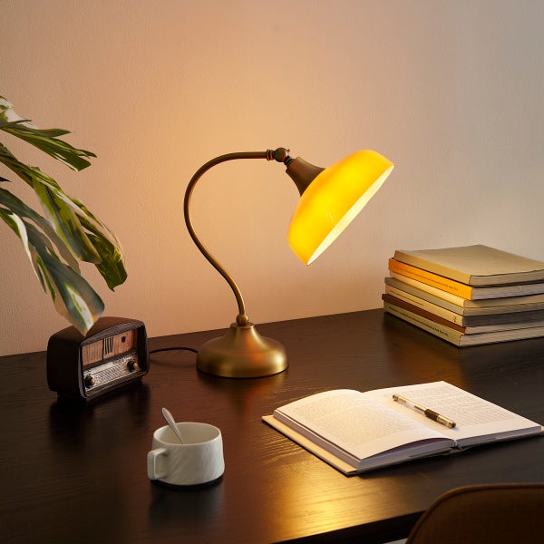 Messing-Schreibtischleuchte mit gelbem Glasschirm auf dunklem Holztisch neben Notizbuch, Büchern und antikem Radio in hell beleuchteter Umgebung.