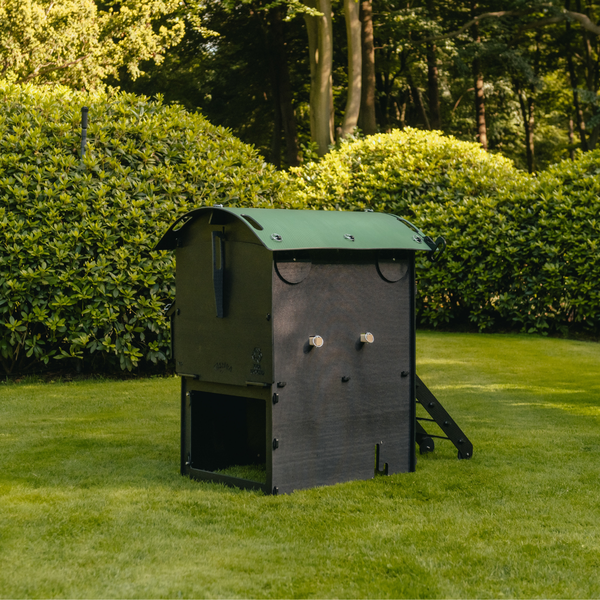 Dunkelgrauer Hühnerstall aus Kunststoff mit grünem Bogendach und Hühnerleiter auf einer Rasenfläche in einem Garten mit Sträuchern.
