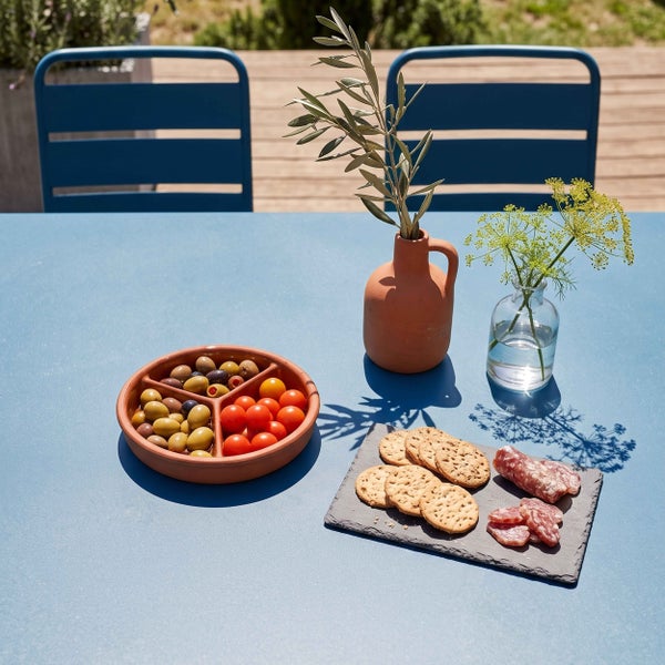 Gedeckter blauer Metall-Gartentisch mit Snacks und Dekoration auf einer Holzterrasse, im Hintergrund zwei passende blaue Gartenstühle.