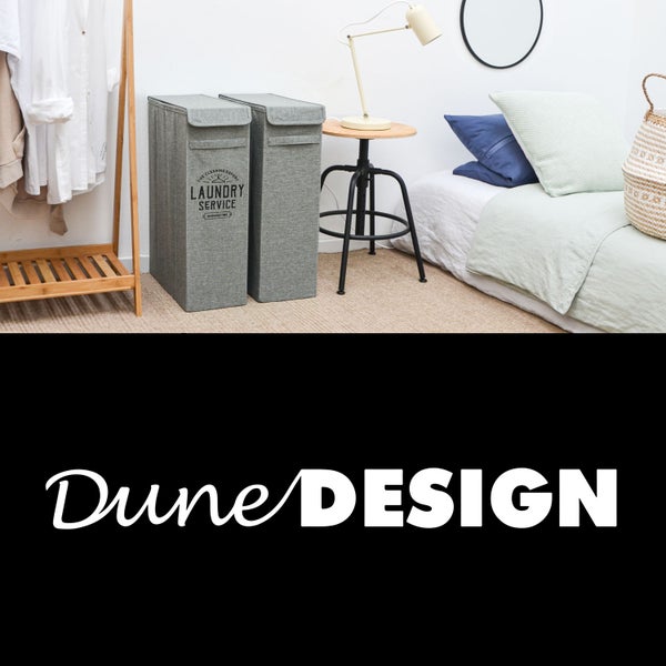 Zwei graue Wäschesammler aus Stoff in einem hellen Schlafzimmer neben einem Beistelltisch und Kleiderständer. DuneDesign Logo.