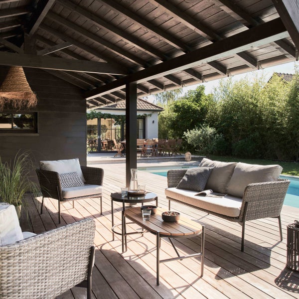 Gartenlounge-Set mit Sofa, Sesseln und Tischen auf einer Holzterrasse unter einem Dach mit Blick auf einen Pool und Garten.