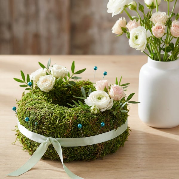 Mooskranz mit Blumen und Dekonadeln neben einer Vase auf Holztisch