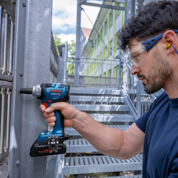 Ein Mann mit Schutzbrille und Gehörschutz bedient einen Bosch Professional Akku-Schlagschrauber an einer Metallkonstruktion im hellen Außenbereich.