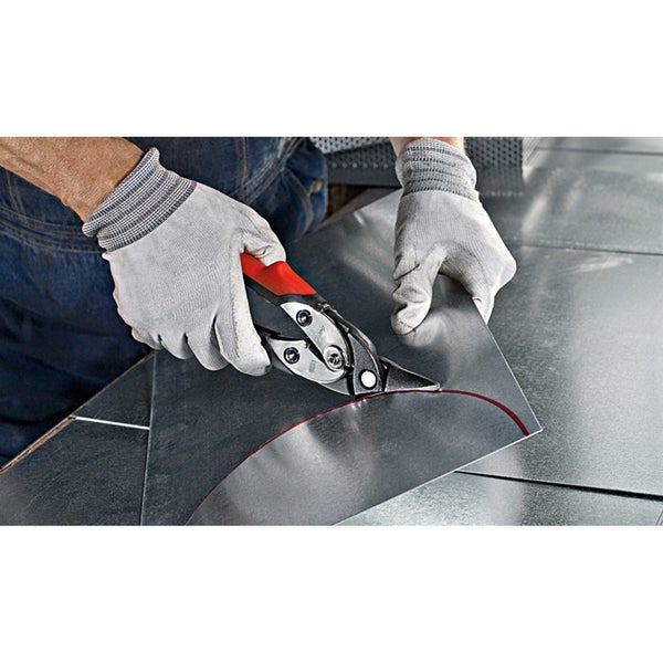 Eine Person mit Arbeitshandschuhen schneidet mit einer Blechschere eine geschwungene Linie in ein Metallblech.