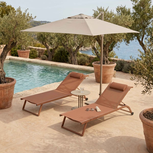 Sonnige Poolterrasse mit zwei braunen Sonnenliegen, Beistelltisch und großem Ampelschirm neben Olivenbäumen in Terrakottatöpfen.
