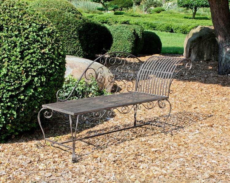 Dekorative Gartenbank aus Metall im antiken Stil mit geschwungenen Ornamenten in einer gepflegten Gartenanlage mit Rindenmulch.