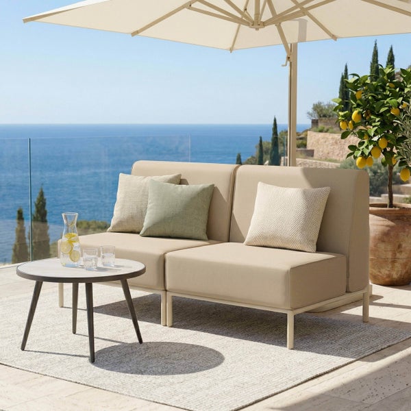 Zweisitzer-Lounge-Sofa mit rundem Beistelltisch unter einem Sonnenschirm auf einer hellen Terrasse mit Meerblick.