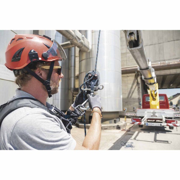 Mann in Industrieumgebung mit Schutzhelm, Visier und Auffanggurt bei der Bedienung eines Seilsicherungssystems mit Karabinerhaken.