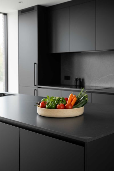 Moderne, minimalistische Küche mit mattschwarzen Fronten, Kochinsel und einer Schale mit frischem Gemüse auf der Arbeitsplatte.