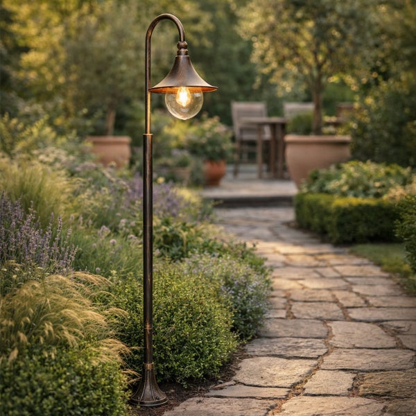 Klassische Außenleuchte aus Bronze an einem gepflasterten Gartenweg, umgeben von Pflanzen und Sträuchern, Sitzgelegenheit im Hintergrund.