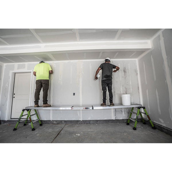 Zwei Arbeiter stehen auf einem Aluminium-Arbeitsgerüst mit zwei Leitern, um eine Trockenbauwand in einem Innenraum zu verspachteln.