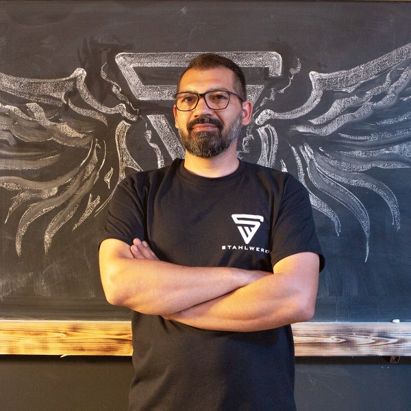 Ein Mann mit verschränkten Armen vor einer Tafel mit dem Stahlwerk Logo und Flügeln.