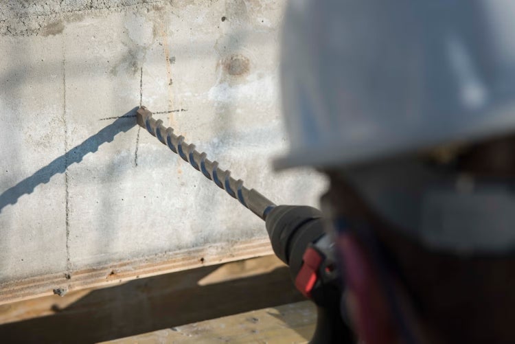Eine Person mit Schutzhelm bohrt mit einem langen Betonbohrer und einer Bohrmaschine in eine Betonwand.