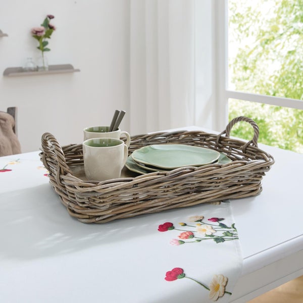 Geflochtenes Weidentablett mit Henkeln auf einem hellen Tisch mit Blumendecke, bestückt mit Keramikgeschirr in einem hellen Raum.