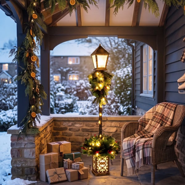 Weihnachtliche Veranda mit einer leuchtenden Laterne, geschmückt mit Tannengrün, Geschenken und einem Korbsessel mit Decke im verschneiten Garten.