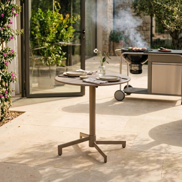 Runder Bistrotisch aus Metall auf einer hellen Steinterrasse, gedeckt für zwei Personen, mit einem modernen Grill im Hintergrund.