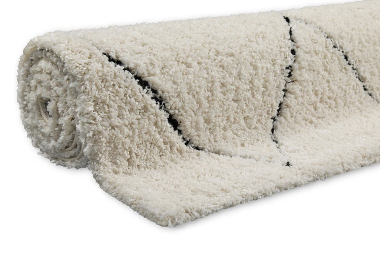 Hochflor-Teppich Shaggy in Cremeweiß mit schwarzem Rautenmuster, teilweise eingerollt, weiche Textur.