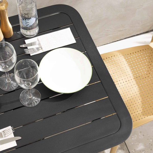 Gedeckter Gartentisch aus schwarzen Metall-Latten mit weißem Teller, Gläsern und Besteck neben einem beigen Stuhl.