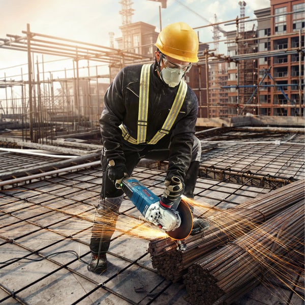 Ein Bauarbeiter mit Schutzhelm und Schutzbrille schneidet mit einem Winkelschleifer Metallstangen auf einer Baustelle.
