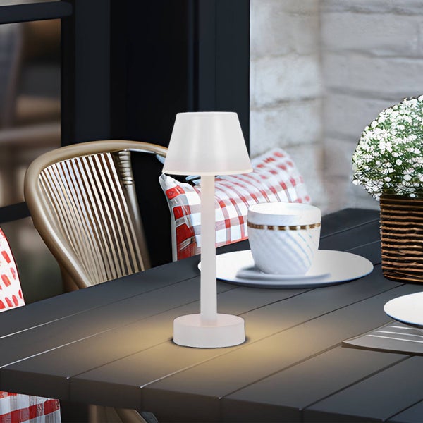 Weiße, kabellose LED-Tischleuchte auf einem dunklen Tisch im Außenbereich, im Hintergrund ein Stuhl und Pflanzen in heller Umgebung.