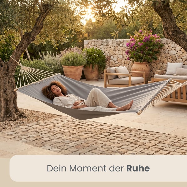 Person in grauer Hängematte auf gepflasterter Terrasse. Im Hintergrund Natursteinmauer, Pflanzen in Terrakotta-Töpfen und Gartenmöbel.