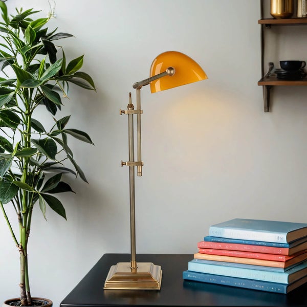 Tischleuchte mit Messingfuß und gelbem Lampenschirm auf einem Tisch neben einem Bücherstapel und einer Pflanze