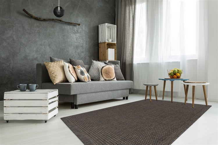Wohnzimmer mit flach gewebtem Teppich vor einem grauen Sofa, dekoriert mit Holzkisten und dekorativen Kissen vor einer strukturierten Wand.