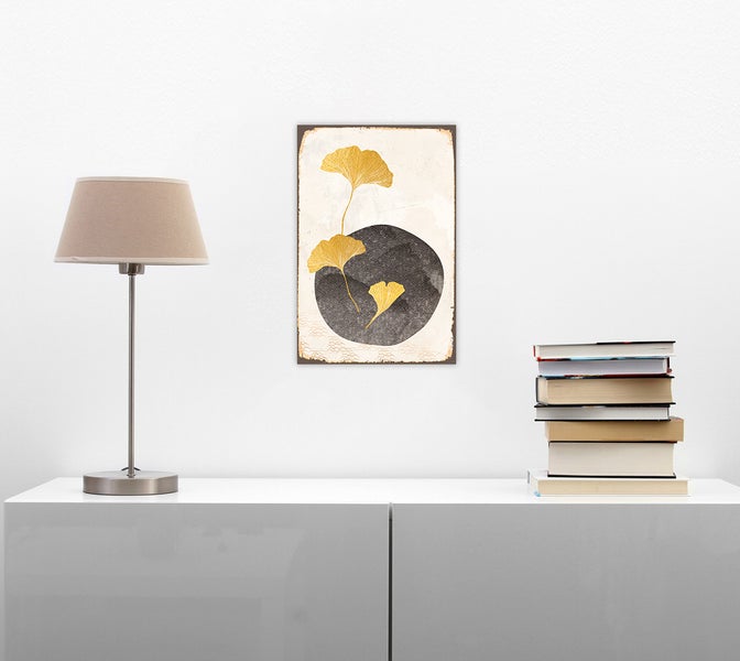 Modernes Wandbild mit goldenen Ginkgoblättern über einem weißen Sideboard mit Tischlampe und Büchern in einem hellen Raum.