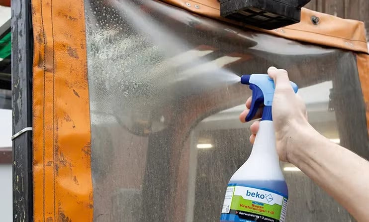 Eine Hand sprüht beko Super Kraftreiniger aus einer Sprühflasche auf eine verschmutzte Kunststoffoberfläche.