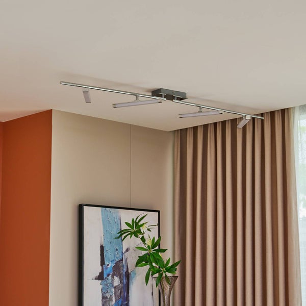 Moderne LED-Deckenleuchte in Chrom-Optik mit vier verstellbaren Strahlern, montiert in einem hellen Wohnzimmer mit Vorhängen und Wandbild.