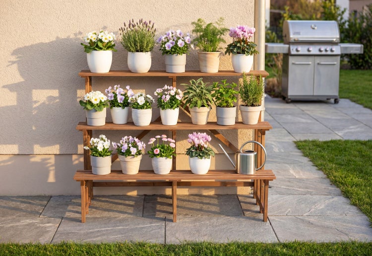 Dreistufiges Pflanzenregal aus Holz auf einer Terrasse mit verschiedenen Blumen und Kräutern in Töpfen sowie einer Gießkanne aus Metall.