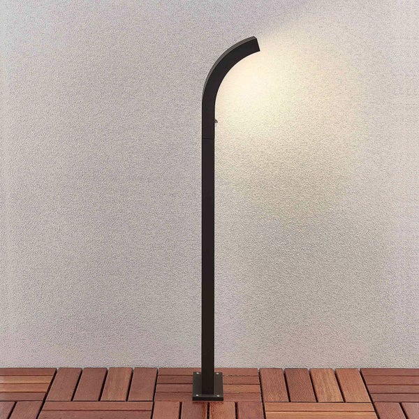 Moderne schwarze LED-Außenstehleuchte mit gebogenem Kopf auf einer Holzterrasse vor einer strukturierten Wand.