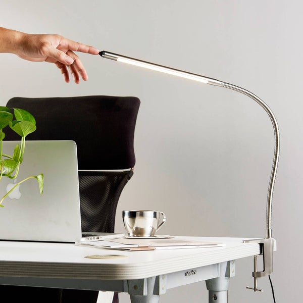 Silberne LED-Klemmleuchte aus Metall mit flexiblem Schwanenhals an einem hellen Arbeitsplatz mit Laptop und Pflanze. Eine Hand berührt die Lampe.