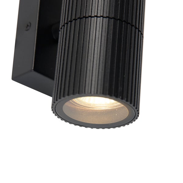 Zylinderförmige Außenwandleuchte aus schwarzem Metall mit geriffelter Oberfläche und eingeschaltetem Licht.