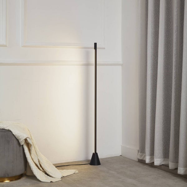 Schwarze LED-Stehleuchte in schlankem Design. Die Lampe strahlt warmweißes Licht gegen eine Wand in einem hellen Raum.
