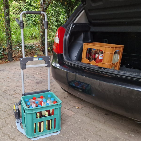 Eine Sackkarre aus Metall steht neben einem Auto und transportiert eine Kunststoffgetränkekiste mit gefüllten Glasflaschen.